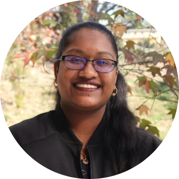 A woman with glasses smiling with foliage in the background.